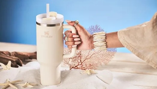 a hand grabbing a Stanley Quencher H2.O FlowState™ Tumbler 40oz. the background is decorated with sand and seashells